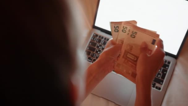 Жінка сидить на ліжку з ноутбуком і рахує євро готівкою — стокове відео