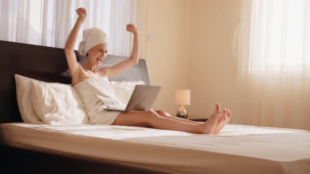 Mientras se acurruca en una acogedora bata de baño y una cama king size, una joven escribiendo en su computadora portátil demuestra que puede trabajar desde cualquier lugar y tener éxito. Mujer usando laptop, sentada en la cama, celebrando el éxito. — Vídeo de stock