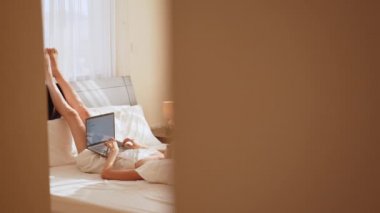 Beyaz yatakta uzanmış gülümseyen bir kadın bacaklarını kaldırırken ve dizüstü bilgisayarda uygulama kullanırken. Güzel genç bir kadının sabah akıllı telefonları kontrol edişinin yüksek açılı görüntüsü. Dizüstü bilgisayar kullanan mutlu kız manzarası.