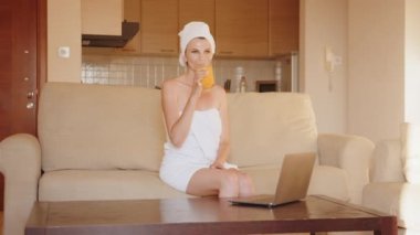 Laptop kullanan ve portakal suyu içen sağlıklı bir kadın. Mutfak masasında duran ve iş arasında portakal suyu içen dizüstü bilgisayarda çalışırken mutlu bir gülümseme.