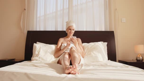 Happy millennial jente som ligger i hvit seng med smarttelefon utstyr ha videosamtale, smilende ung kvinne slappe av i soverom våkne opp i morgen tekstmeldinger på celle en enhet, teknologi konsept – stockvideo