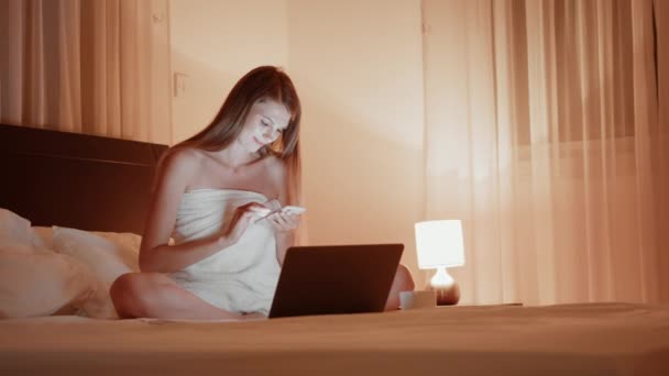Dizüstü bilgisayarı ve akıllı telefonuyla yatakta oturan gülümseyen kadın. — Stok video
