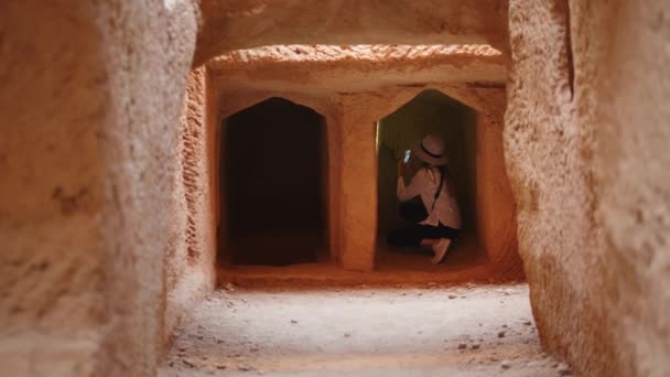 Mujer caucásica en sombrero blanco con linterna en un teléfono inteligente moderno mientras examina pasajes subterráneos de templo viejo. Expedición arqueológica sobre la civilización antigua. Mujer examinando templo antiguo — Vídeos de Stock