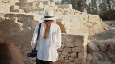 Antik sütunları inceleyen kadın arkeolog. Fotoğraf makinesi ve kalıntılar arasında antik tapınak keşfeden notları olan beyaz bir kadın. Yazlık şapkalı kadın yıkılmış tapınağın arasında yürüyor.