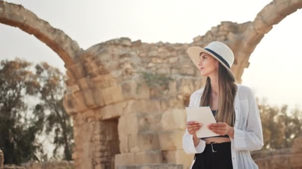 Жінка в білому капелюсі і повсякденному одязі, що ходить серед зруйнованого стародавнього храму з картою в руках. Археолог відкриває покинуті історичні місця. Археолог досліджує старі історичні будівлі — стокове відео
