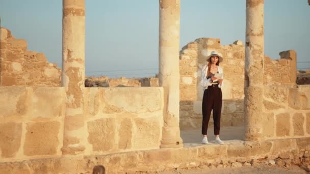 Girl Tourist In an ancient temple with columns looking at records of a map. Competent female archeologist standing among ruins of ancient temple. Woman holding brush for digging and map in hands. — Vídeo de Stock