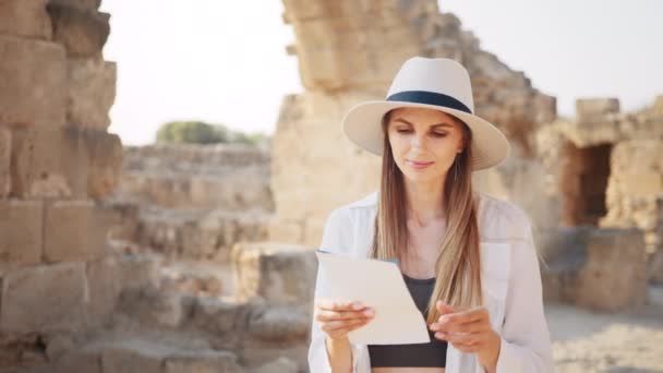 Vrouw met witte hoed en casual kleding wandelen tussen ruïneuze oude tempel met kaart in handen. Archeoloog ontdekt verlaten historische plaatsen. Archeoloog onderzoekt oude historische gebouwen. — Stockvideo