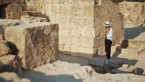 美丽的女人带着笔记和照相相机在被毁的古寺上进行挖掘。戴着白帽的考古学家花了夏天的时间来发现历史珍品.古建筑的残破 — 图库视频影像
