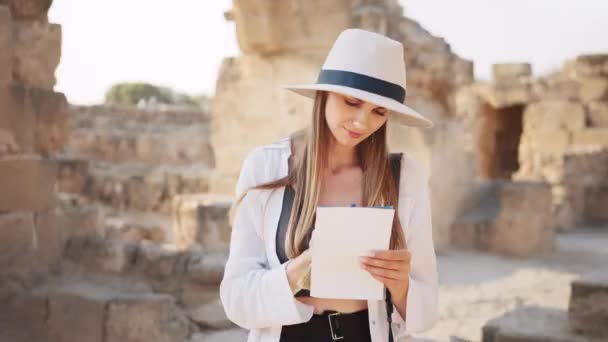 Woman among ruined ancient temple with map in hands. Archeologist discovering abandoned historical places. Archeologist examining old historical buildings. Girl researcher makes notes — Vídeo de Stock