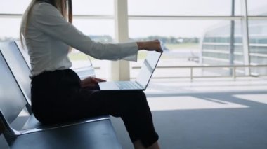 Havaalanında dizüstü bilgisayarda oturan genç ve güzel bir kadın. Kız turist serbest çalışıyor ve bekleme odasında uçmayı bekliyor. Konsept seyahat, uzaktan çalışma. Laptop kullanan bir bayanla havaalanı salonu.