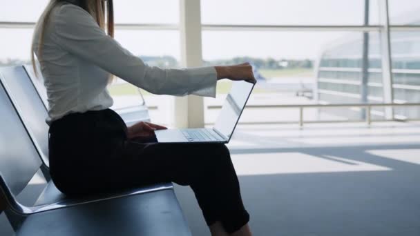 Young pretty woman printing on laptop sitting at airport. Girl tourist freelancer works and waits for flight in waiting room. Concept travel, remote work. Airport lounge with a lady using a laptop — Stock Video