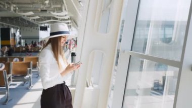 Kız havaalanı salonunda akıllı telefon kullanıyor. Akıllı telefon iletişim uygulamasını kullanan güzel genç bir kız yeni bir ülkeye geliyor. Kadınlar dünyayı dolaşır. Kameraya gülümsüyor.