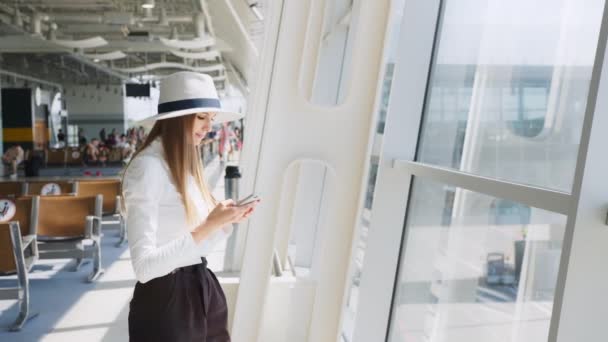 Κορίτσι χρησιμοποιεί smartphone στο σαλόνι του αεροδρομίου. Όμορφη νεαρή κοπέλα που χρησιμοποιεί το αεροδρόμιο εφαρμογή smartphone επικοινωνίας που φθάνουν στη νέα χώρα. Γυναίκα ταξιδεύει σε όλο τον κόσμο. Χαμογελώντας στην κάμερα — Αρχείο Βίντεο