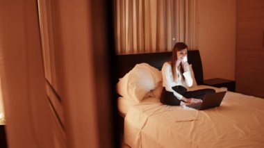 İş gezisi sırasında otel odasında yatarken cep telefonuyla konuşan ve bilgisayarla iletişim kuran pozitif ırklı bir kadın. Çekici bir iş kadını ailesiyle uzaktan iletişim kuruyor.