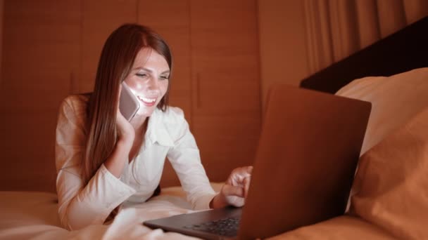 Kafkasyalı genç bir kadın rahat bir yatakta yatarken dizüstü bilgisayarla konuşuyor ve daktilo kullanıyor. İş gezisi sırasında otel odasında yatakta oturan bir kadın cep telefonuyla ve bilgisayarla konuşuyor.. — Stok video