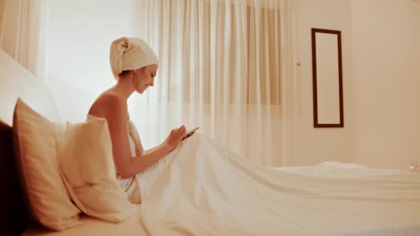 Πλευρική άποψη της καυκασίας γυναίκα γραπτών μηνυμάτων στο σύγχρονο κινητό τηλέφωνο, ενώ στηρίζεται σε άνετο κρεβάτι του δωματίου του ξενοδοχείου. Όμορφη κυρία τυλιγμένη σε πετσέτα μπάνιου χρησιμοποιώντας smartphone κατά τη διάρκεια των διακοπών. Γυναίκα που χρησιμοποιεί κινητό — Αρχείο Βίντεο