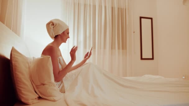 Güzellik merkezindeki güzellik prosedürleri. Havlu sarıklı, beyaz elbiseli mutlu kadın otel odasında cep telefonuyla konuşuyor. Kadın rahat bir yatakta dinlenirken cep telefonuyla mesajlaşıyor. — Stok video