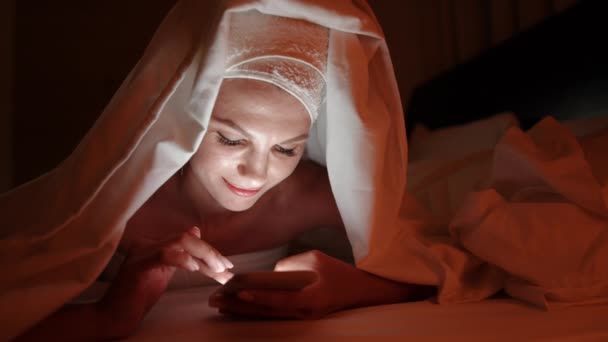 Yatak odasında battaniyenin altında akıllı telefon kullanan güzel bir genç kadın. Kadınlar akıllı telefon kullanır ve geceleri yatakta yatarlar. İnsanlar, uyku zamanı ve dinlenme konsepti - yatakta akıllı telefonlu genç bir kadın — Stok video