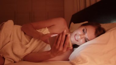 Yatakta cep telefonuyla mesajlaşan genç bir kadın. Yatak odasında cep telefonu olan yorgun bir kız. Gece yatakta uzanmış telefona bakan bir kadın. Uyumadan önce mavi cep telefonu ekranına bakan kişi.