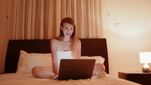 Angenehme junge Frau lächelt und winkt beim Videoanruf auf modernem Laptop. Glückliche Dame im Badetuch mit Online-Kommunikation am Abend im Hotelzimmer. — Stockvideo