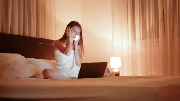 Close up of happy positive Caucasian young beautiful woman calling on cellphone while resting laying on bed in bedroom. Cozy hotel room with evening light. Smiling woman sitting on bed with smartphone — 图库视频影像