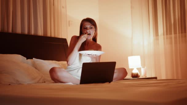 Happy lady watching movie and eating tasty meal during evening time at hotel room. Pretty woman in bath towel using wireless laptop on comfy bed. Woman watching film and eating dish at hotel room — 图库视频影像