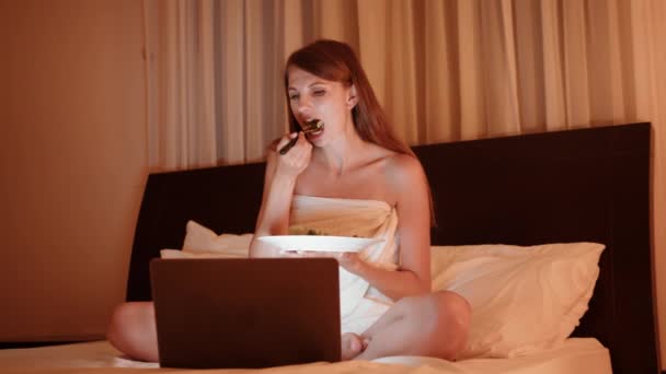 Молода жінка сидить на ліжку з тарілкою смачної страви і дивиться фільм на сучасному ноутбуці. Щаслива сім'я насолоджується вечерею в затишному готельному номері і використовує комп'ютер. Жінка п'є сік і дивиться фільм — стокове відео