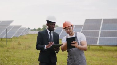 Dışarıda güneş panellerini inceleyen çok ırklı iki adam. Takım elbiseli Afrikalı Amerikalı işadamı ve panoya yazı yazarken dijital tablet kullanan Hintli teknisyenle konuşurken.