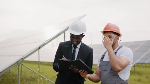 技术员和检查员站在太阳能发电厂中间.身穿黄色硬帽和安全眼镜的印度工程师手持数码平板电脑，与太阳能发电厂的男性所有者交谈。能源的核查. — 图库视频影像