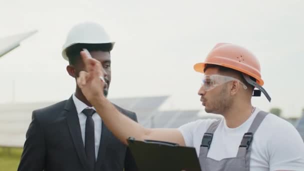 Close up of african man in suit signing documents on clipboard that holding indian engineer. Two multiracial people standing among solar farm and making agreement. Men and technician among solar cells — Stock Video