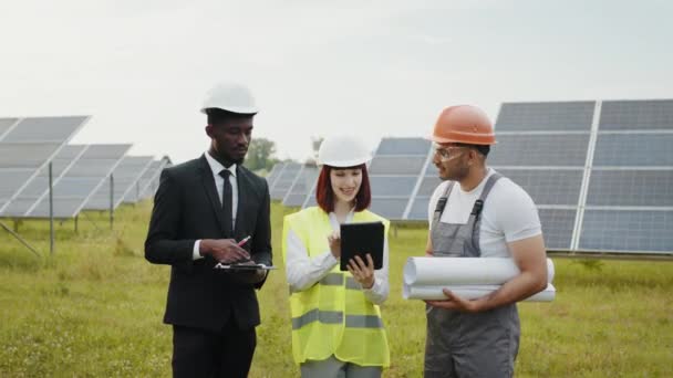 Gente multiétnica en cascos posando en una granja solar. Mujer caucásica y hombre africano de pie con el ingeniero indio entre los paneles solares y mirando a la cámara. Técnico de inspección de personas en granja solar — Vídeos de Stock