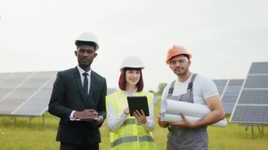 Kafkas kadın ve Afrikalı adam güneş panelleri arasında Hint mühendisiyle dikilip kameraya bakıyor. Dijital tablet, pano ve çizimler tutan insanlar. Çok kültürlü insanlar güneş çiftliğinde çalışıyor.