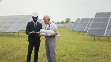 Miğferli Hintli teknisyen iki müfettişle güneş santralinde duruyor, gülümsüyor ve bakıyor. Erkek ve kadın mühendisler iş için pano ve tablet kullanıyorlar. Çok kültürlü insanlar güneş çiftliğinde çalışıyor.
