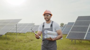 Üniformalı Hintli mühendis portresi, sarı kask ve güneş çiftliğinde dururken elinde planlar ve pano tutan güvenlik gözlükleri. İnsanlar ve alternatif enerji kavramı. Güneş çiftliğinde mühendis.