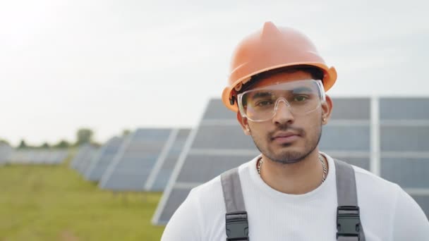 Ingeniero en la granja solar. Retrato de ingeniero indio en uniforme, casco amarillo y gafas de seguridad sosteniendo planos y portapapeles mientras está parado en la granja solar. Concepto de personas y energía alternativa — Vídeos de Stock