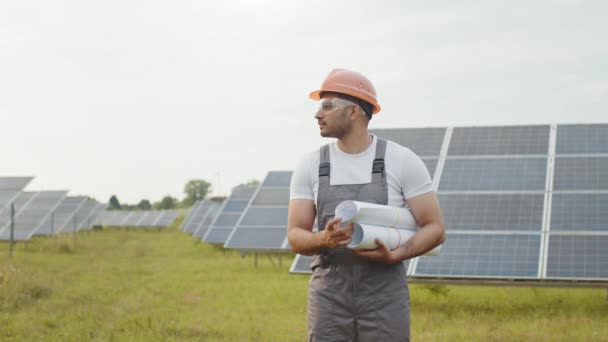 Técnico indio sonriente en casco amarillo y gafas de seguridad posando en la estación solar. Trabajador sosteniendo planos en las manos. Producción de energía verde. Técnico en casco posando en estación solar — Vídeos de Stock