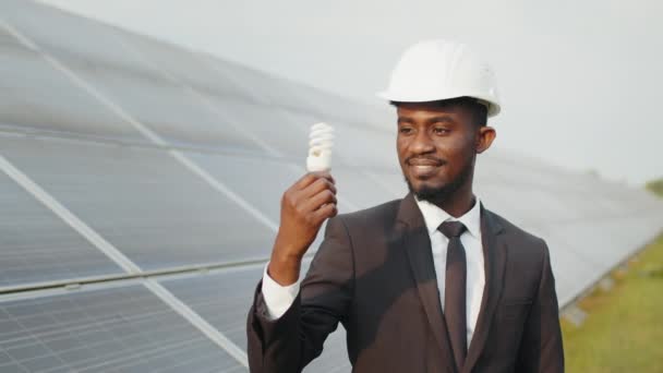 手に低エネルギー電球とカメラに笑みを浮かべて白いハード帽子と黒のスーツでアフリカ系アメリカ人の男性の肖像画。ソーラーパネル付きの駅の背景。電球のついた太陽光発電所のアフリカ人 — ストック動画