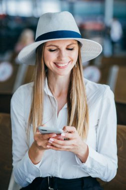 Tablet akıllı telefon kullanarak seyahat et. Genç iş kadını gülümseyerek seyahat valiziyle havaalanındaki bekleme salonunda oturuyor..