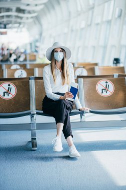 Maskeli genç bir kadın salgın sırasında bir havaalanı salonunda oturur ve pasaportunu ve kamera biletini gösterir.