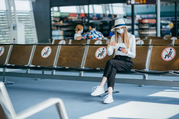 Kobieta w wieku 25-35 lat, ubrana w kapelusz i maskę ochronną, siedzi przy kasie odpraw w terminalu lotniska, czeka na odlot, trzyma bilety lotnicze z powodu podróży. — Zdjęcie stockowe