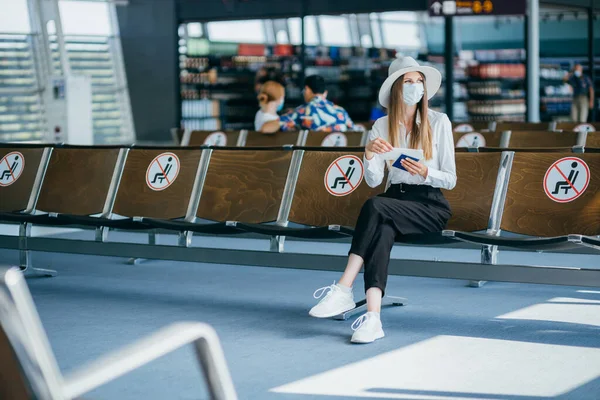 Die selbstbewusste Geschäftsfrau in Maske, die am Flughafen sitzt, einen Pass und Flugtickets in der Hand hält, hält bei einer Pandemie Abstand. Warten am Flughafen. Reisekonzept. Frau schaut sich Fahrkarten an. — Stockfoto