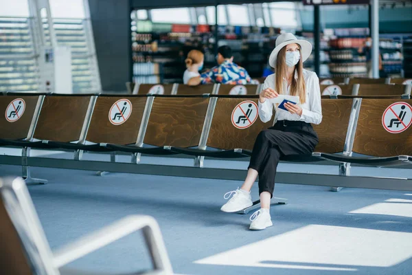 Turisti maski istuu lentokentän terminaalin lounge tyhjiä merkittyjä tuoleja uusien normaalien ja covid sosiaalisen etäisyyden määräyksiä. Nainen istuu lentokentän loungessa ja katselee passia ja matkustaa — kuvapankkivalokuva