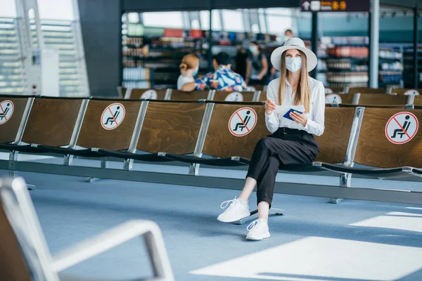 Nuori nainen turisti yllään kasvonaamio tilalla passi ja lippu, istuu lentokentällä tai rautatieasemalla tyhjällä merkitty tuolit uusien normaalin ja sosiaalisen covid etäisyyden asetusten — kuvapankkivalokuva