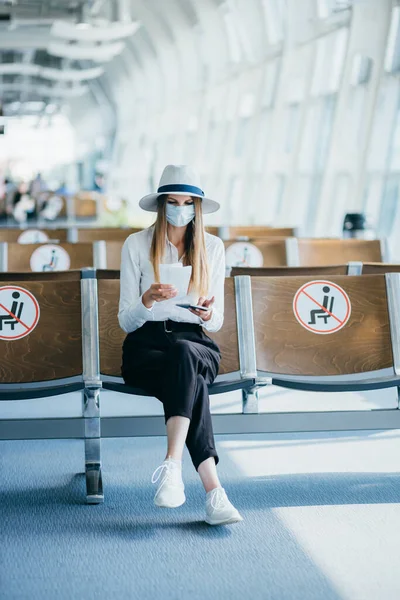 Die selbstbewusste Geschäftsfrau in Maske, die am Flughafen sitzt, einen Pass und Flugtickets in der Hand hält, hält bei einer Pandemie Abstand. Warten am Flughafen. Reisekonzept. Frau schaut sich Fahrkarten an — Stockfoto