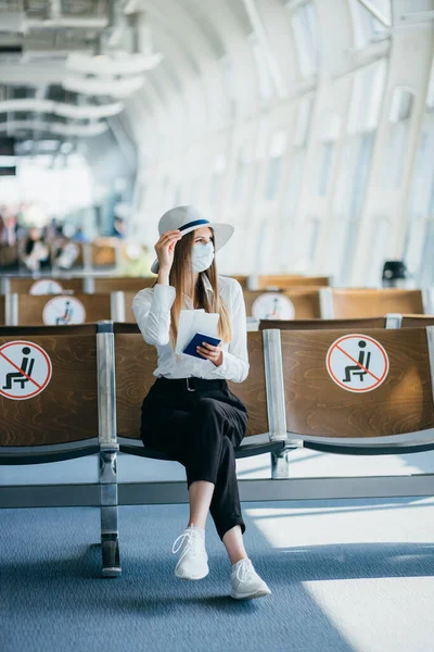 Nuori nainen turisti yllään kasvonaamio tilalla passi ja lippu ja käyttämällä älypuhelin, istuu lentokentällä tai rautatieasemalla tyhjällä merkitty tuolit uuden normaalin ja sosiaalisen covid etäisyyden sääntelyn — kuvapankkivalokuva