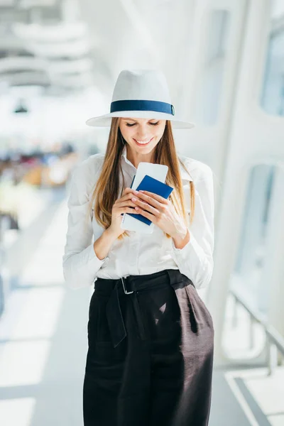 Piękna bizneswoman na lotnisku, używająca telefonu do wysyłania SMS-ów, stojąca przy oknie w biurowcu korporacyjnym, sprawdzająca wiadomości. Dziewczyna trzyma bilety i paszport.. — Zdjęcie stockowe