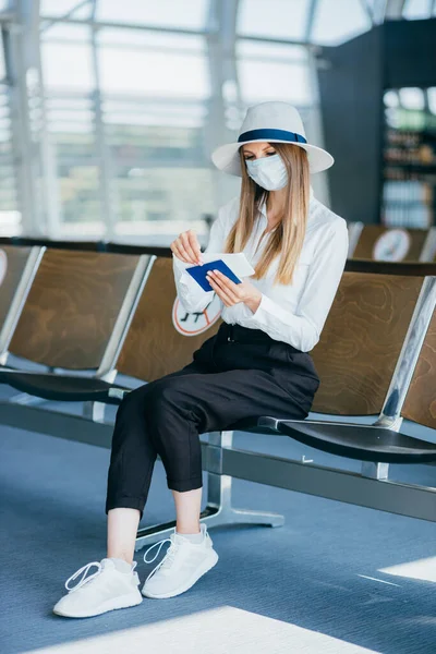 Junge Frau mit medizinischem Mundschutz steht vor einer Informationstafel am Flughafen oder Bahnhof. Frauen mit Reisepass und Flugticket und Reisen während der Pandemie. Stockbild