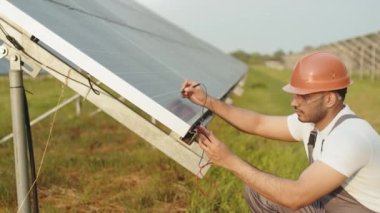 Güvenlik kaskı ve güneş panellerinin amperini çoklu metreyle ölçen uzman teknisyen. Hintli adam dışarıda fotovoltaik hücreleri onarıyor. Yeşil enerji konsepti. Güneş panellerinin genişliği