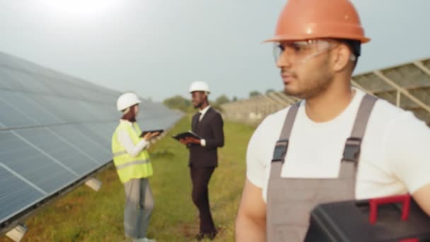 Indian man in overalls, glasses and orange helmet holding instruments and looking aside while standing among solar station. Group of engineers having discuss on background. Technician at solar farm — Stock Video