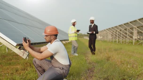 Tecnico energetico in casco e uniforme utilizzando multimetro per misurare la tensione dei pannelli solari all'aperto. Gruppo di ingegneri con tablet e appunti in piedi su sfondo. Uomo vicino pannello solare — Video Stock